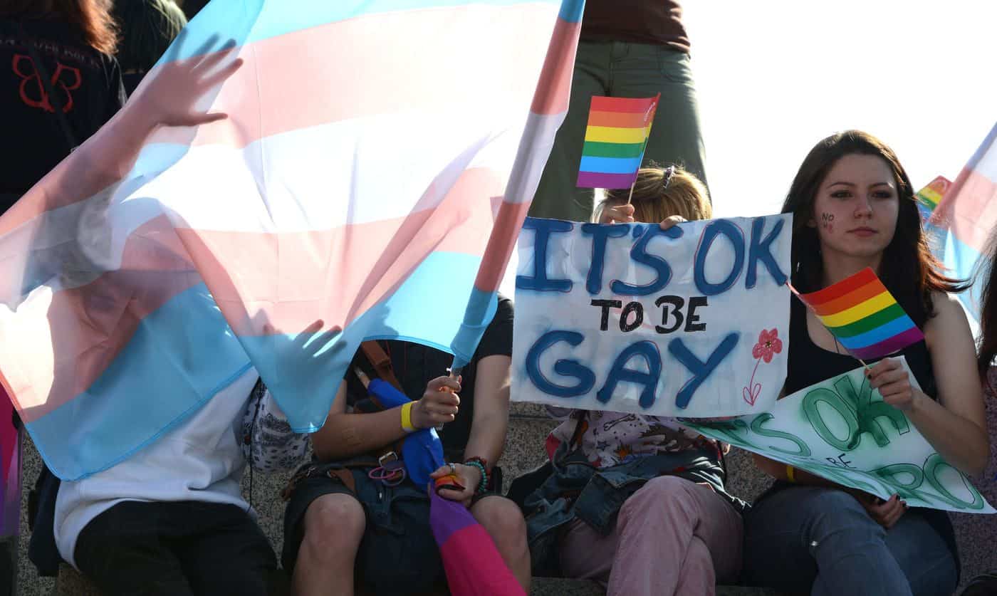 Bandera trans y cartel con mensaje "It’s OK to be gay" en manifestación LGBTQ+.