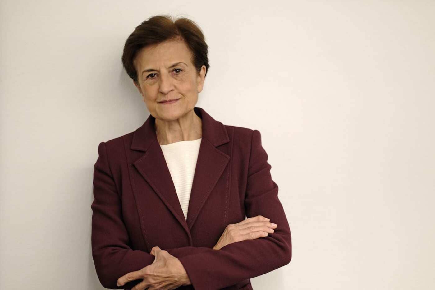 Pensadora mayor en fotografía, mujer con abrigo burdeos, estilo serio, fondo blanco, expresión confiada, plataforma de divulgación filosófica.