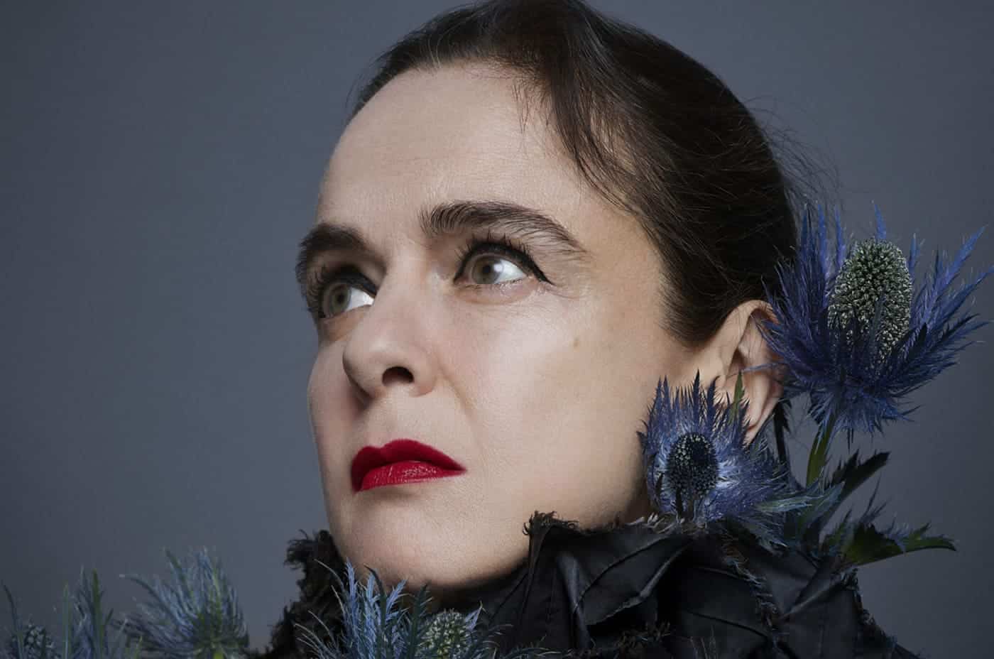 Profesora con flores azules en el cabello y expresión pensativa en fondo gris.