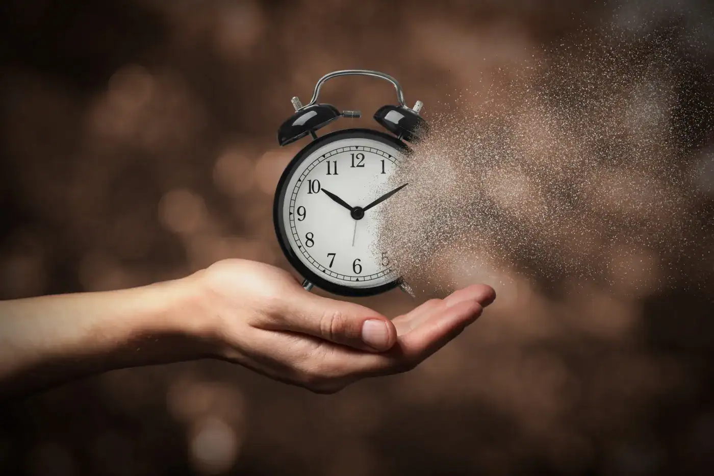 Reloj despertador en mano disipándose en polvo en un fondo borroso.