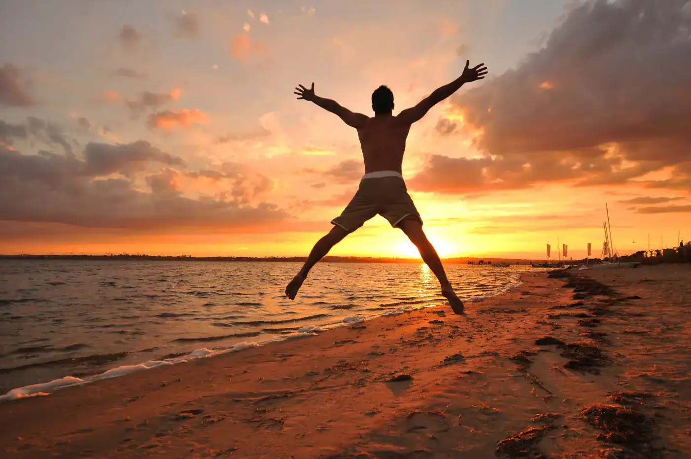 Varios pasos de pies en la playa durante el atardecer con cielo nublado y tonos anaranjados, y hombre saltando con los brazos abiertos.