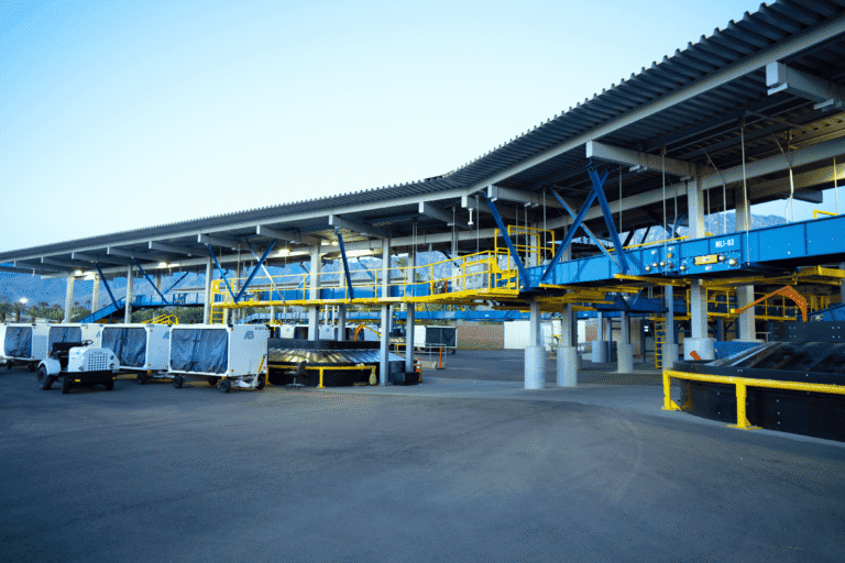 An outdoor baggage handling facility with blue and yellow conveyor systems, white baggage carts, and a tug vehicle under a large canopy, with mountains in the background.