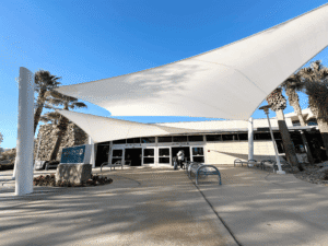 Entrance to a building with white sail-like shade structures, palm trees, and a sign for "GATES 12 TO 20" under a clear blue sky.