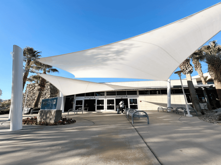 Entrance to a building with white sail-like shade structures, palm trees, and a sign for "GATES 12 TO 20" under a clear blue sky.