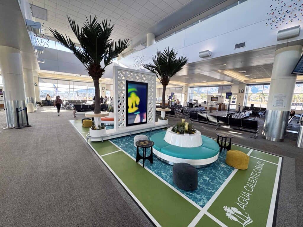 A modern airport lounge with a green and blue patterned floor, circular turquoise seating, artificial palm trees, and a large digital art screen. Standard airport seating and windows looking out to a tarmac and mountains are in the background.