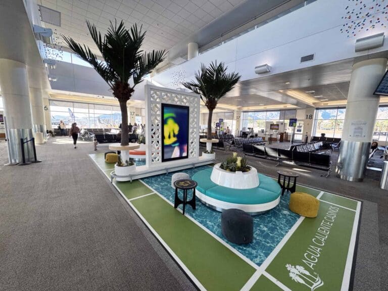 A modern airport lounge with a green and blue patterned floor, circular turquoise seating, artificial palm trees, and a large digital art screen. Standard airport seating and windows looking out to a tarmac and mountains are in the background.