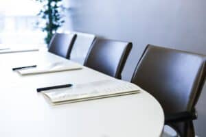 A conference table with documents and pens, and several dark chairs in a bright room.