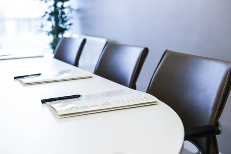 A conference table with documents and pens, and several dark chairs in a bright room.