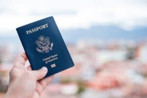 A hand holds a dark blue United States passport, showing its front cover with gold lettering and the Great Seal, against a blurred outdoor background.