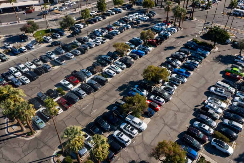 Aerial view of a large, crowded parking lot with hundreds of cars, SUVs, and trucks, surrounded by palm trees.