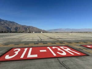 Airport runway with '31L-13R' markings in red and white, an air traffic control tower, and rugged mountains under a clear blue sky.