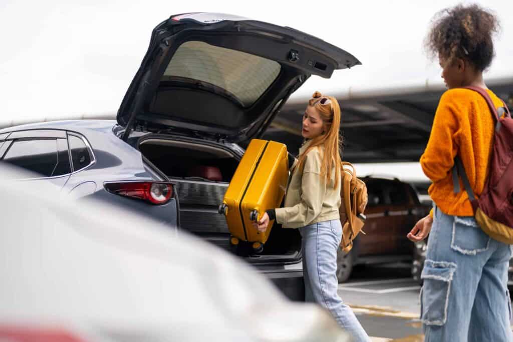 A young woman loads a yellow suitcase into a car trunk, with another person standing nearby.