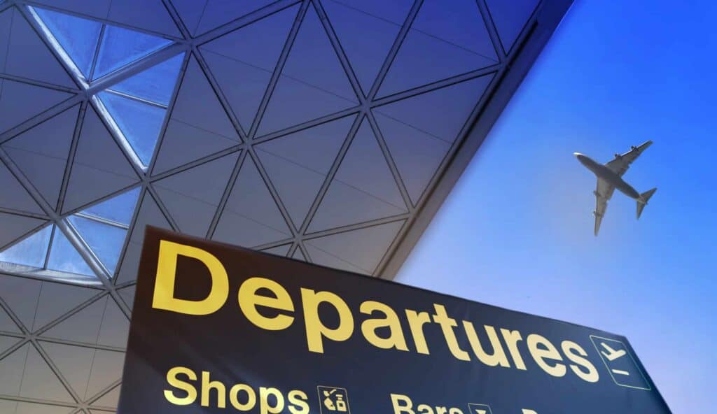 A black 'Departures' sign with yellow text in an airport, with a modern geometric ceiling and an airplane flying in a blue sky above.