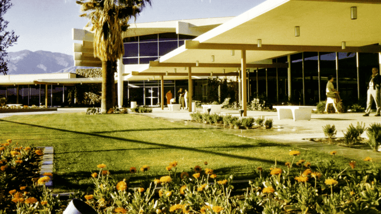 A mid-century modern building with a flat, overhanging roof, large windows, and a stone facade, surrounded by a green lawn, flowers, and a palm tree. Mountains are visible in the background.