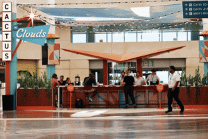 A modern bar named 'Cactus to Clouds' with patrons, staff, cacti planters, and string lights, located in what appears to be an airport terminal.