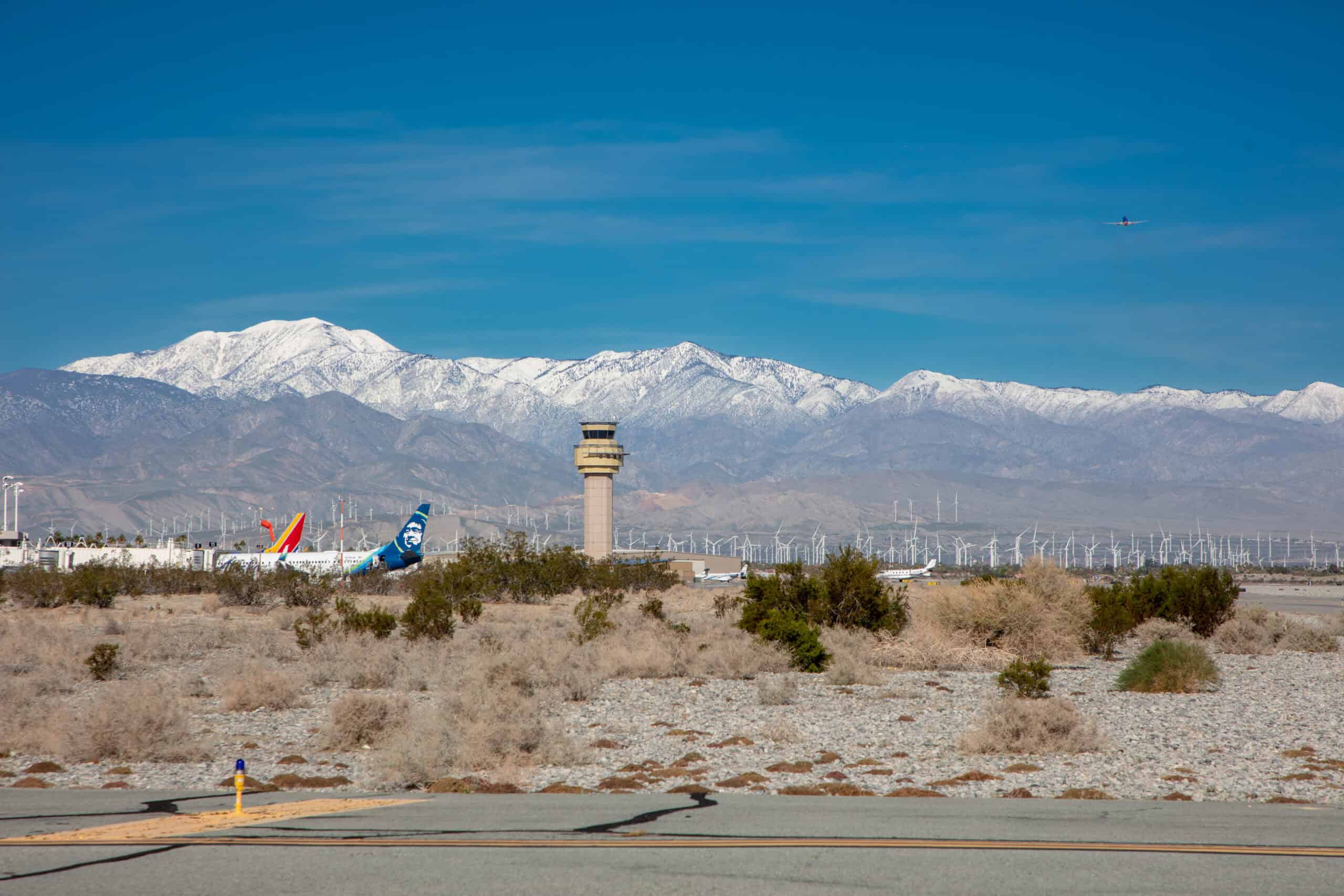 An airport with a control tower and parked planes, including an Alaska Airlines jet, set against a backdrop of a wind farm and snow-capped mountains under a clear blue sky. A small plane flies in the distance.