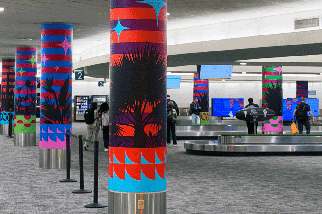 Airport baggage claim area with large, colorful pillars featuring palm tree and tropical plant designs, alongside people waiting for luggage at carousels.