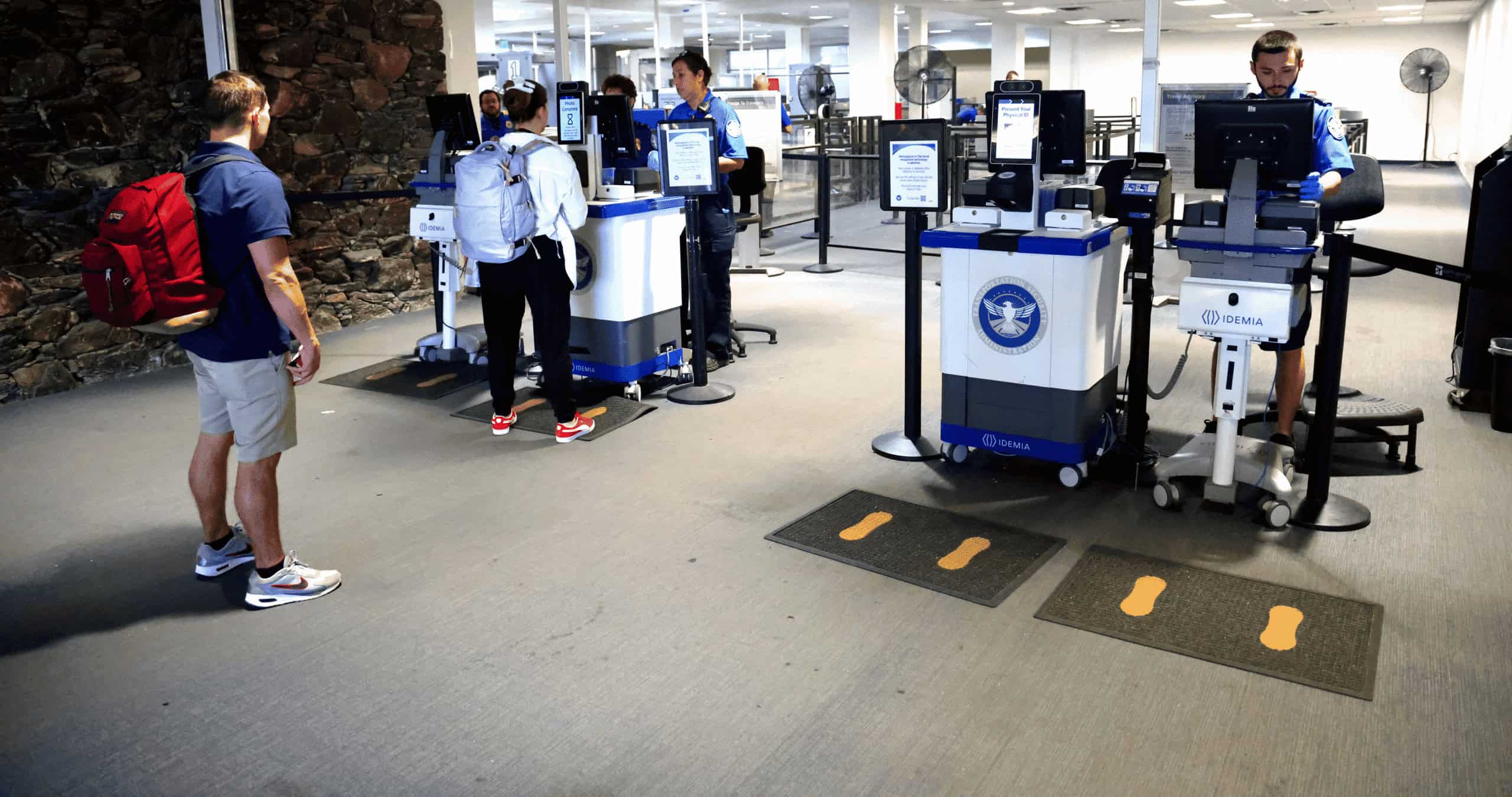 People using self-service kiosks at an airport security checkpoint, with uniformed staff present.
