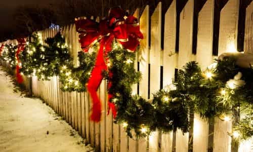 A white picket fence decorated with green garlands, red bows, and eco-friendly light decorations glows warmly during winter at night.