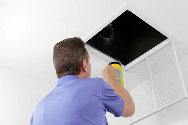 A man in a blue shirt shines a flashlight into an open attic access panel in a white ceiling, inspecting for Air Duct Repair needs in Vernon, BC.