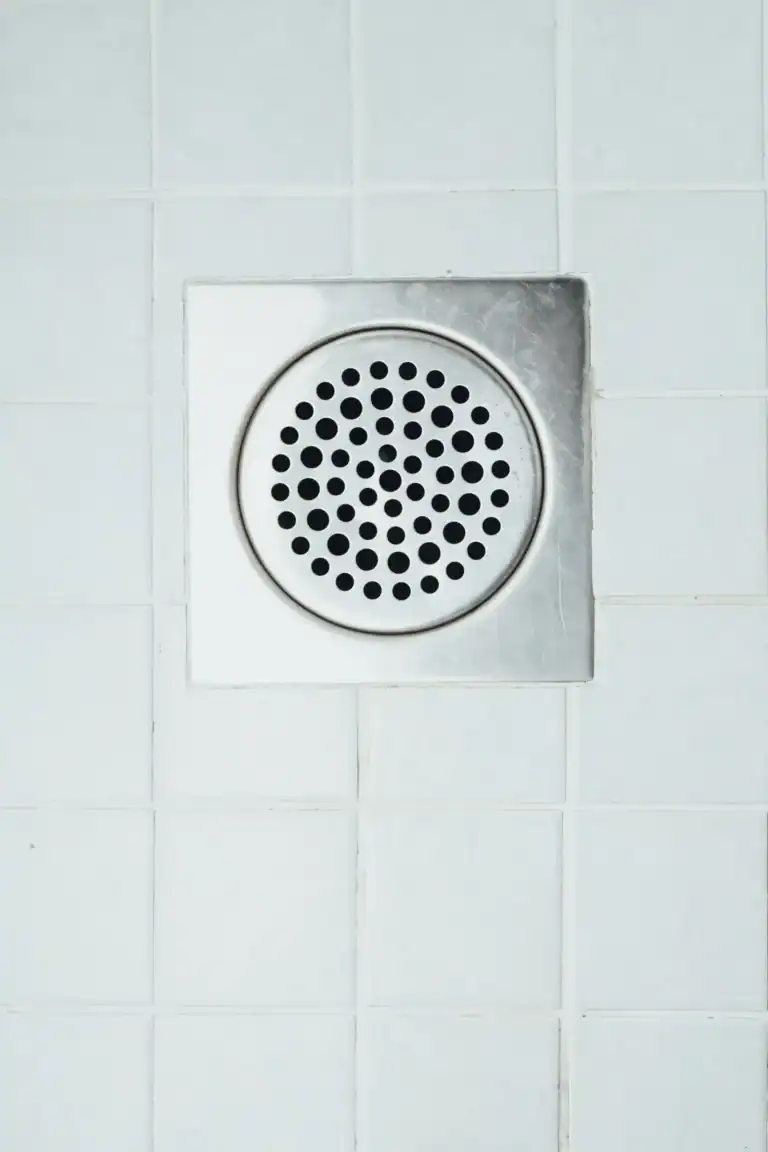 A metal shower drain cover with circular holes is set into white tiled flooring, ensuring easy access for Rooter Service when needed.