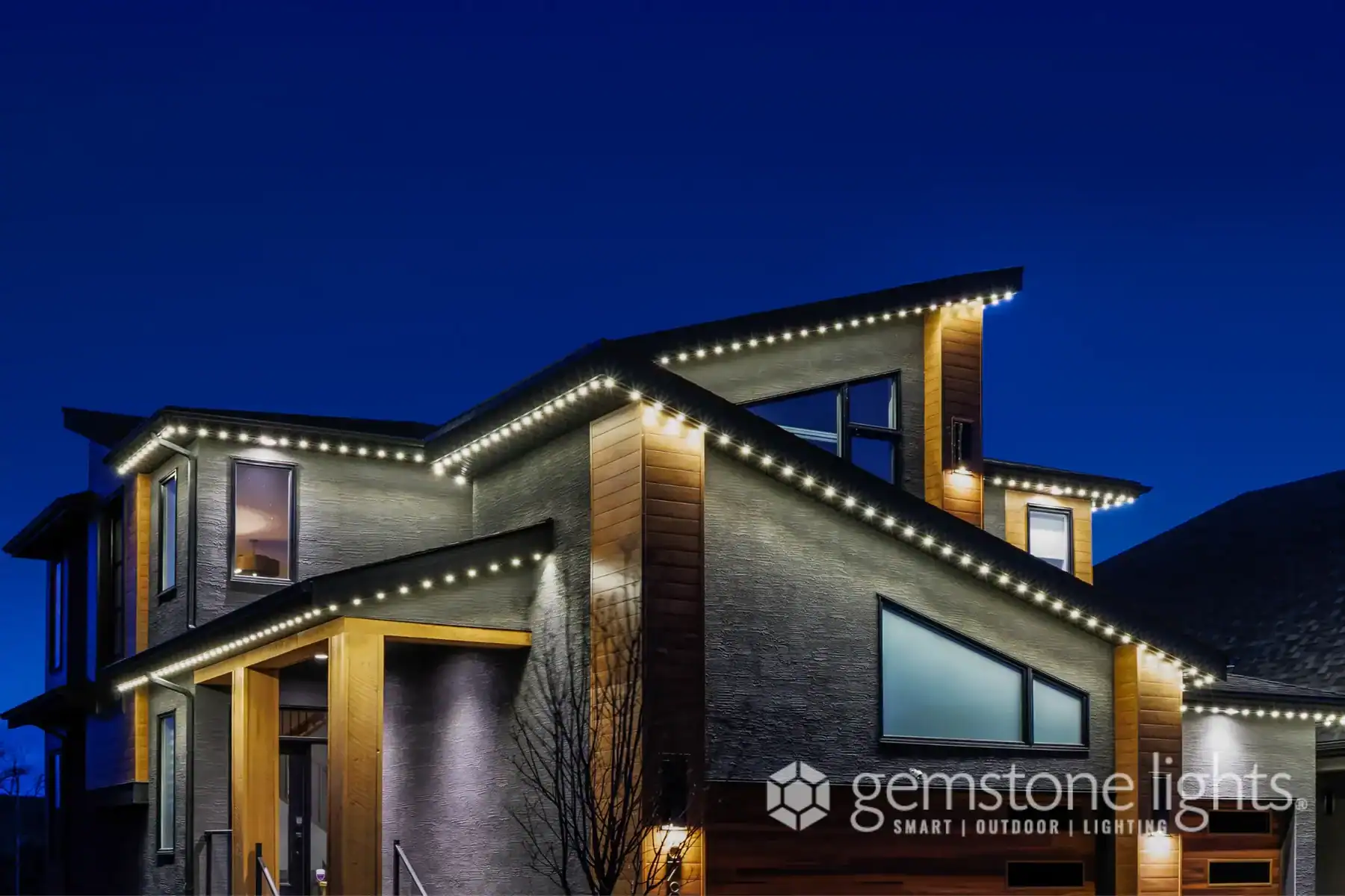 Modern house at night with LED trim lighting along the roof edges, featuring wood and gray accents, and large windows; “gemstone lights” logo in corner—discover more about us and our innovative lighting solutions.
