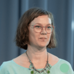 Inga Schiffler lächelt eine Kamera. Sie hat kinnlange, braune Haare, eine Brille, ein grünes Oberteil und eine farblich passende Kette mit grünen Steinen.