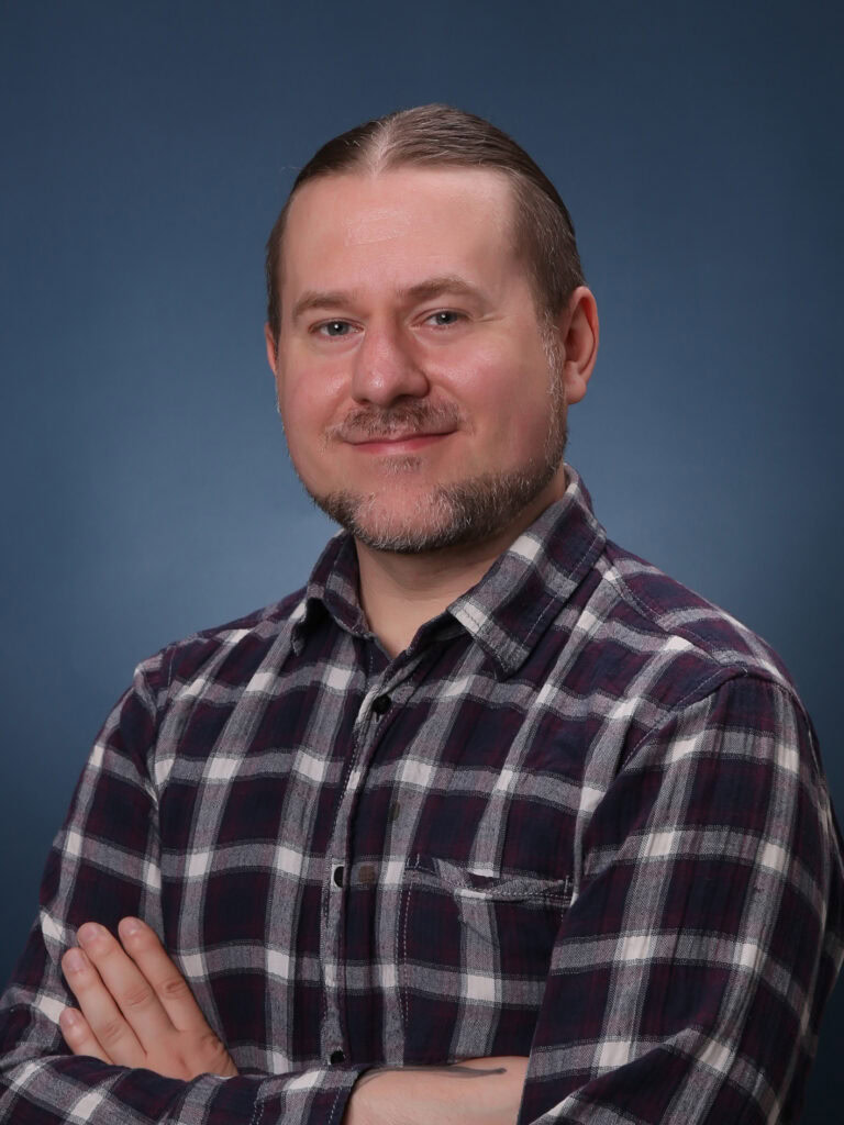 Profilfoto von Sascha Bach, er lächelt in die Kamera und trägt ein blau kariertes Holzfällerhemd.