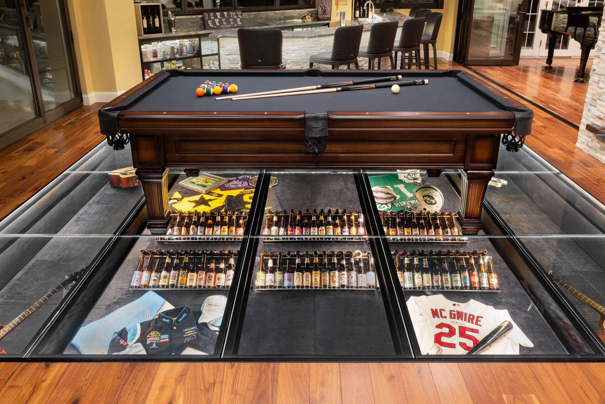Pool table on a walkable glass floor displaying beer bottles, sports uniforms and guitars encased beneath tempered glass