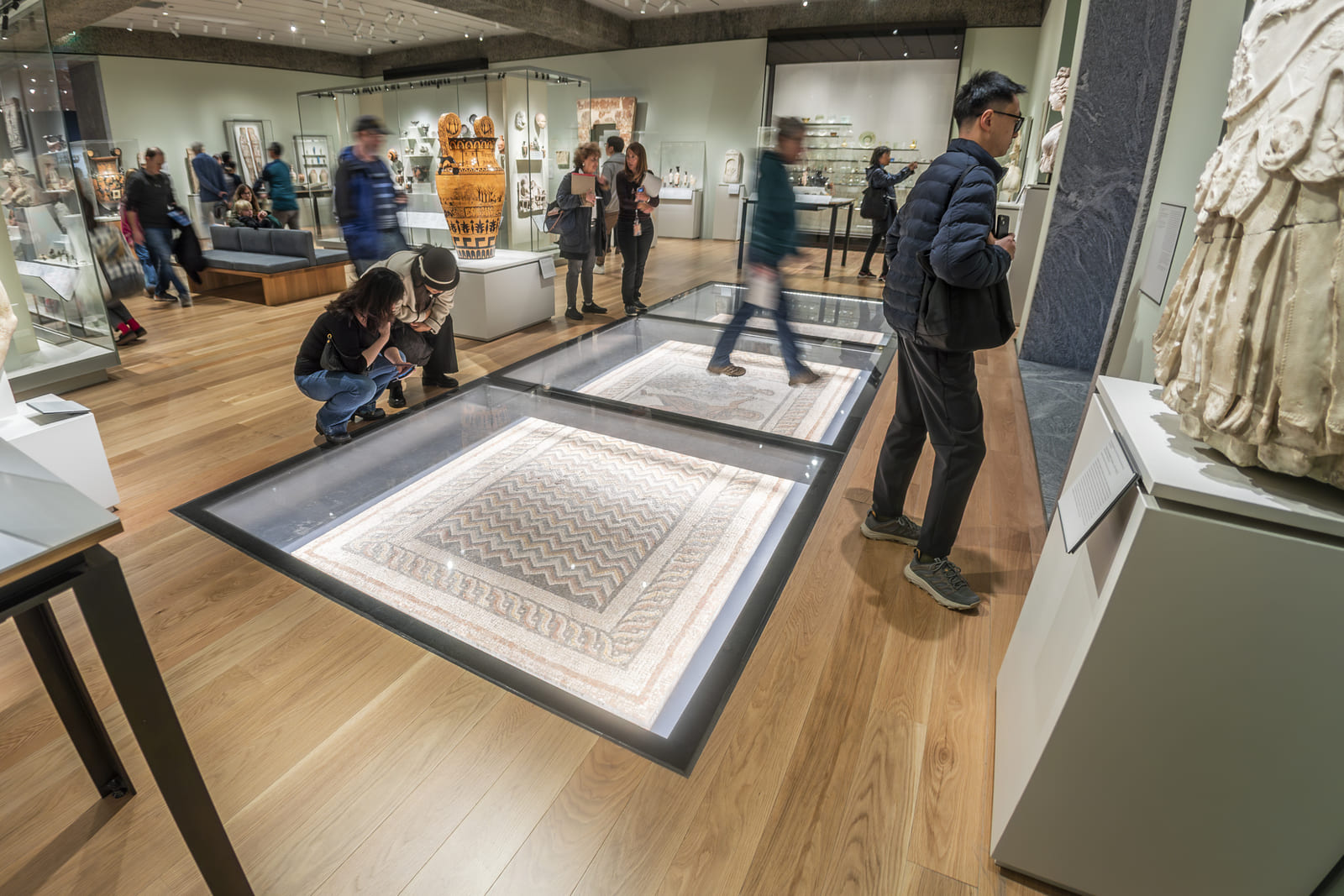Museum visitors around a large illuminated glass floor display case set into a wooden gallery floor