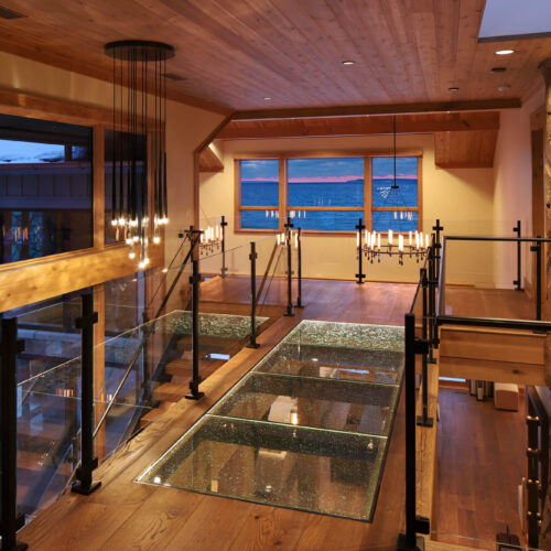 Upper-level structural glass floor bridge with glass railings in a timber lakefront home at dusk, indoor climbing wall at right