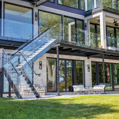 Rear of a modern lakefront home with a structural glass deck, glass railings and an exterior glass staircase descending to the lawn