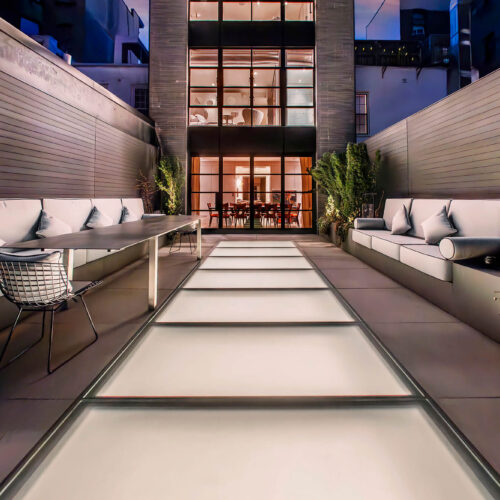 Rooftop terrace at night with a flush SkyFloor Series 2000 walkable skylight glowing between built-in seating, townhouse windows lit behind