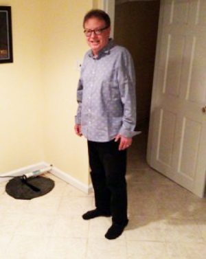 A man wearing glasses, a light blue button-up shirt, black pants, and black socks stands and smiles in a room with light-colored walls and tiled floor.
