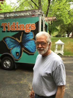 An older man with gray hair and glasses stands outside near a truck with a large blue butterfly and the word "Tidings" on its side. Trees and a yard are in the background.