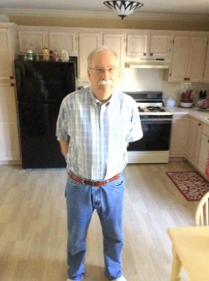 An older man with gray hair and a mustache stands in a kitchen with light wood cabinets, a black refrigerator, and a stove in the background.