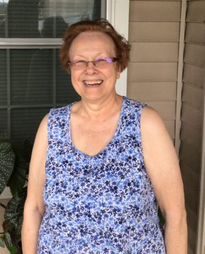 An older woman wearing glasses and a blue floral sleeveless top stands smiling in front of a window and beige siding.