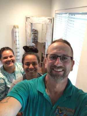 Three people smiling indoors, with a man in glasses taking a selfie, two women standing behind him, and a window with blinds in the background.