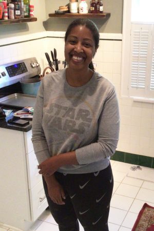 A woman standing and smiling in a kitchen, wearing a gray Star Wars sweatshirt and black pants with a white Nike logo. Shelves with jars and a stove are visible in the background.