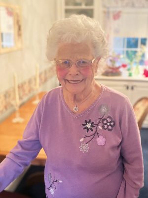 An elderly woman with white hair and glasses smiles while standing in a dining room, wearing a lavender sweater with floral embroidery.