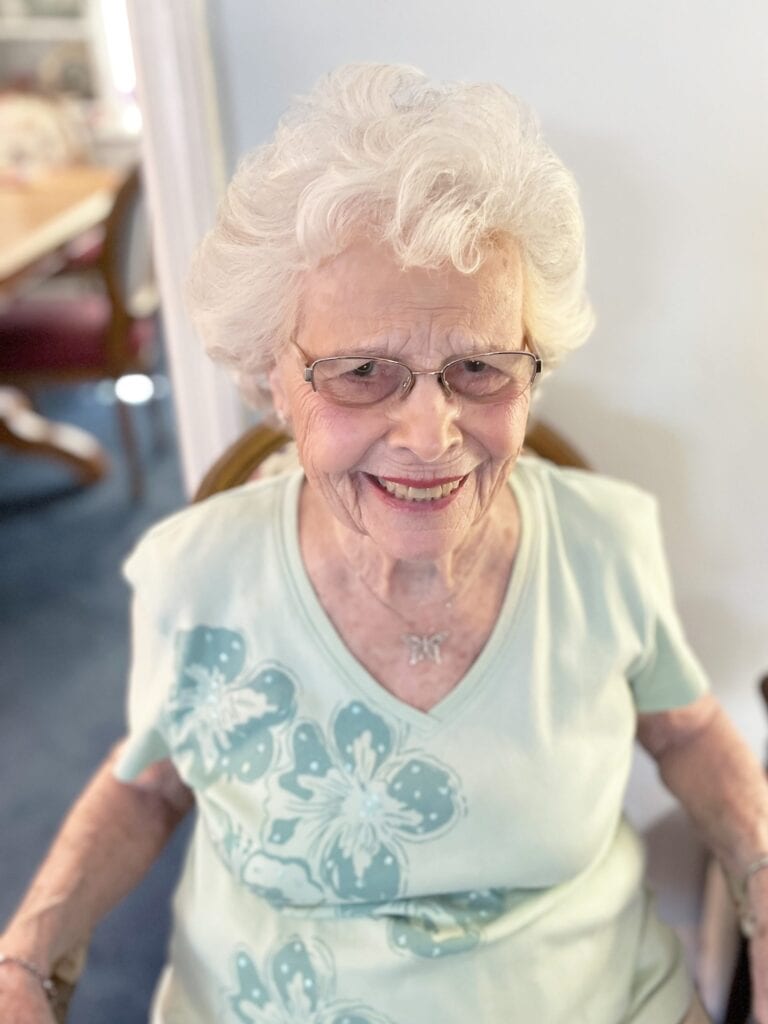 Elderly woman with white hair, glasses, and a light blue floral shirt smiles while sitting indoors.