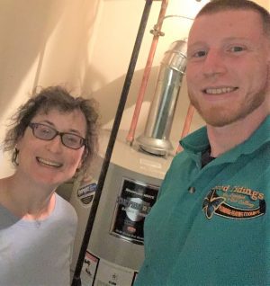 Two people standing and smiling in front of a water heater; one person is wearing a blue company shirt.