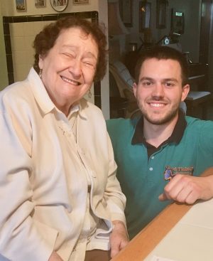 An older woman and a younger man sit side by side at a kitchen counter, both smiling at the camera.