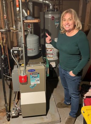 A woman stands in a basement next to a furnace and water heater, giving a thumbs-up.
