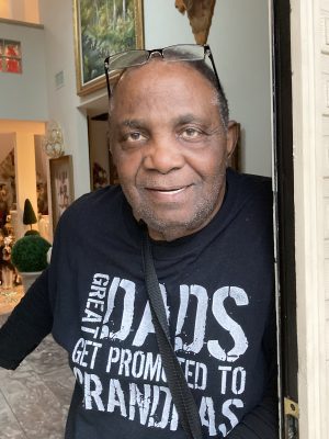 An older man stands in a doorway, smiling. He wears glasses on his head and a black shirt that says, "Great Dads Get Promoted to Grandpas.