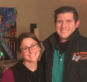 Two people standing indoors, smiling at the camera; one is wearing glasses, and the other is wearing a jacket with embroidered text on it.