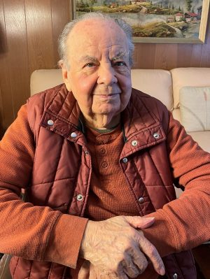 An elderly man sits on a light-colored couch, wearing an orange shirt and a maroon quilted vest, with his hands resting in his lap. A painting hangs on the wood-paneled wall behind him.