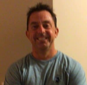 A man with short dark hair wearing a light blue shirt is smiling in front of a beige and white wall.