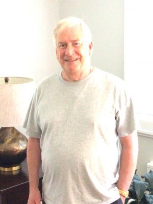 An older man with short white hair, wearing a light gray t-shirt, stands indoors near a lamp and window, smiling at the camera.