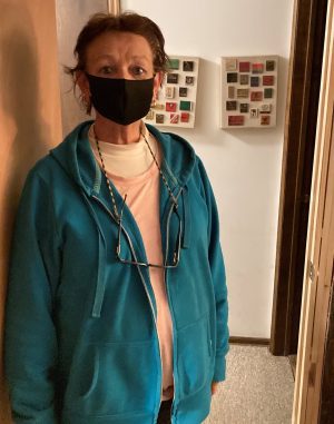 Woman wearing a black face mask, teal zip-up hoodie, and glasses around her neck, standing in a hallway with framed displays on the wall behind her.